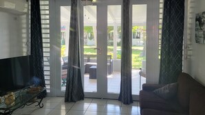A living room with a sofa, a glass coffee table, and a TV. There is a sliding glass door leading to an outdoor patio area with furniture.