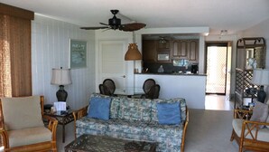 A living room with a sofa, chairs, a coffee table, a ceiling fan, and a kitchen in the background.
