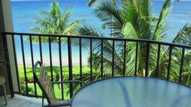 A balcony with a glass table and chairs overlooking a beach with palm trees and the ocean.