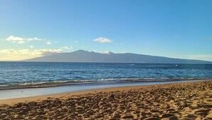 A beach with footprints leading to the water, a person relaxing on the sand, and a mountain in the background.