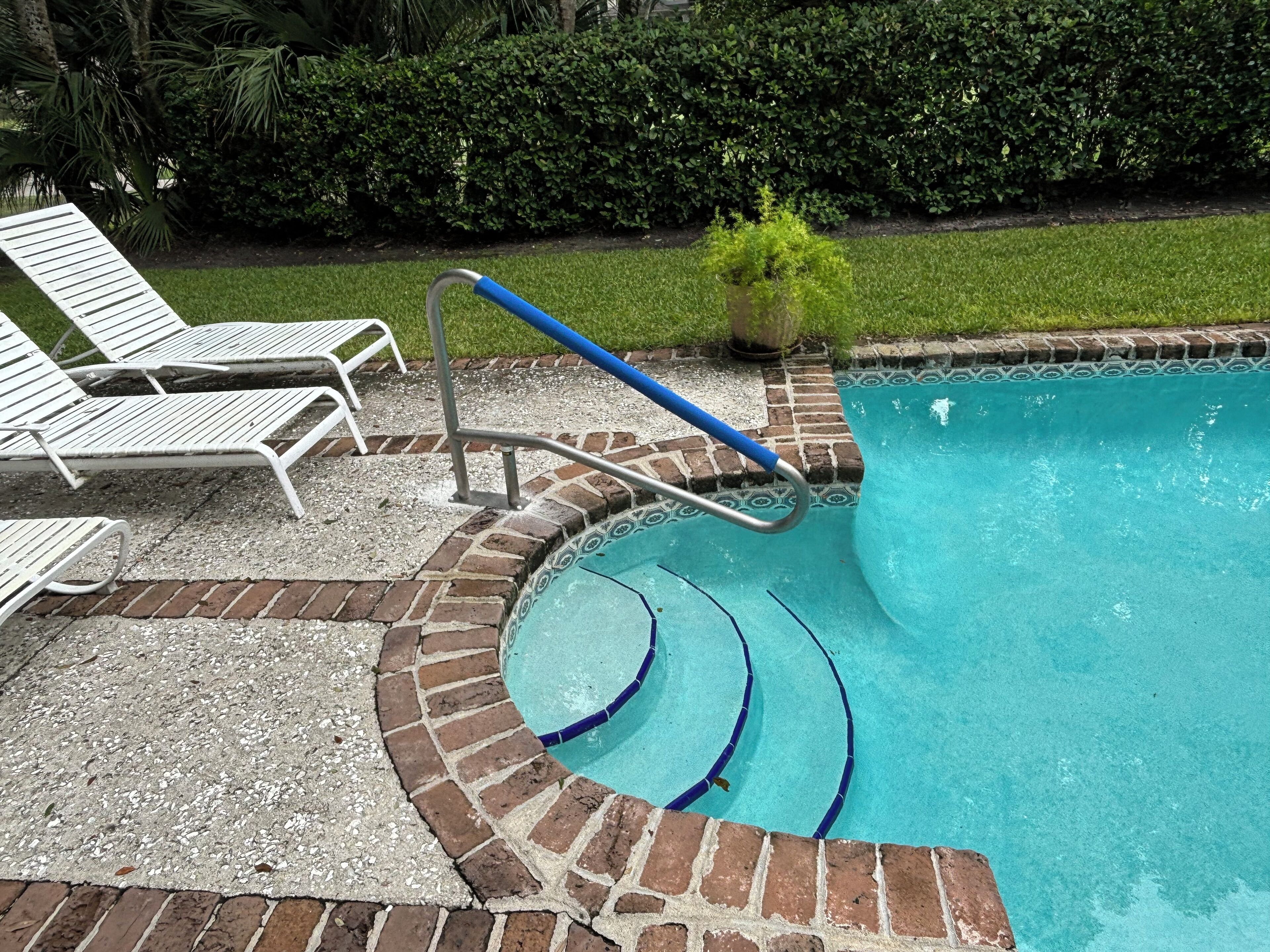 Una piscina con borde de ladrillo, barandilla azul y sillas reclinables blancas.