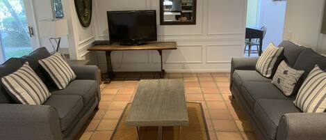 Una sala de estar con dos sofás grises, una mesa de centro, un televisor sobre un mueble de madera y un piso de baldosas.