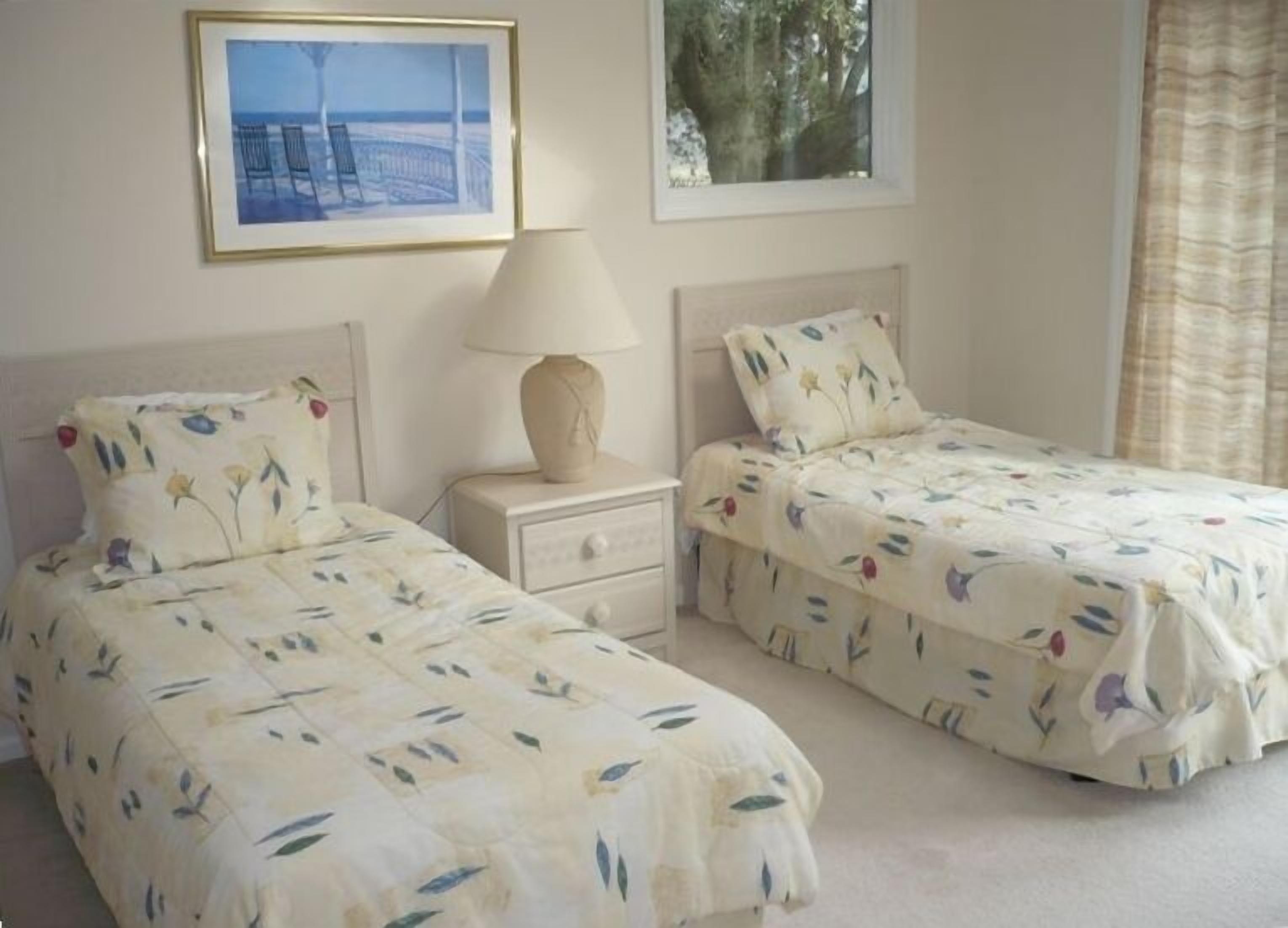 Un dormitorio con dos camas individuales, una mesita de noche con lámpara y un cuadro enmarcado en la pared.