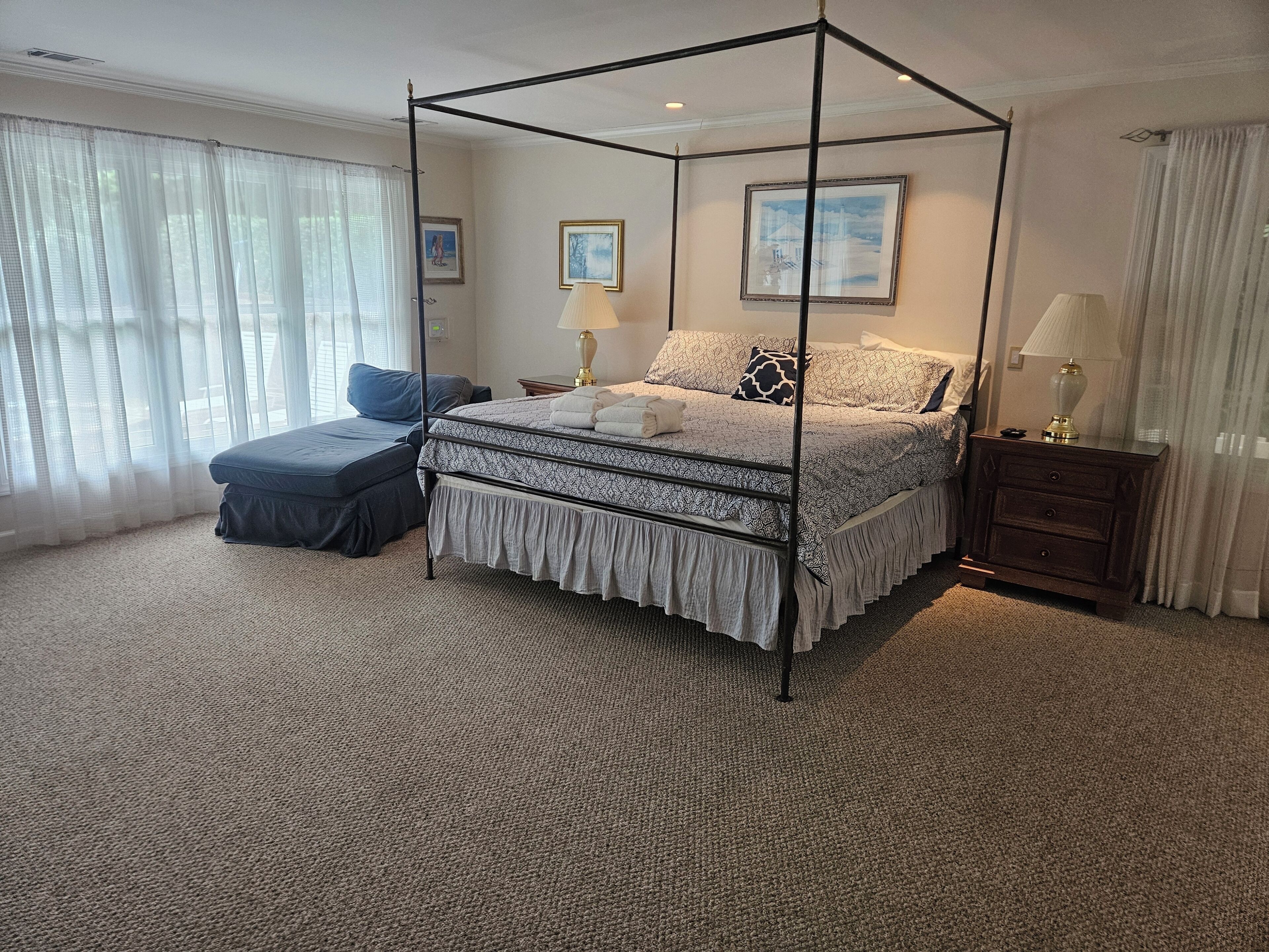 A four-poster bed with a ruffled skirt, a footstool, a nightstand with a lamp, and a framed picture on the wall.