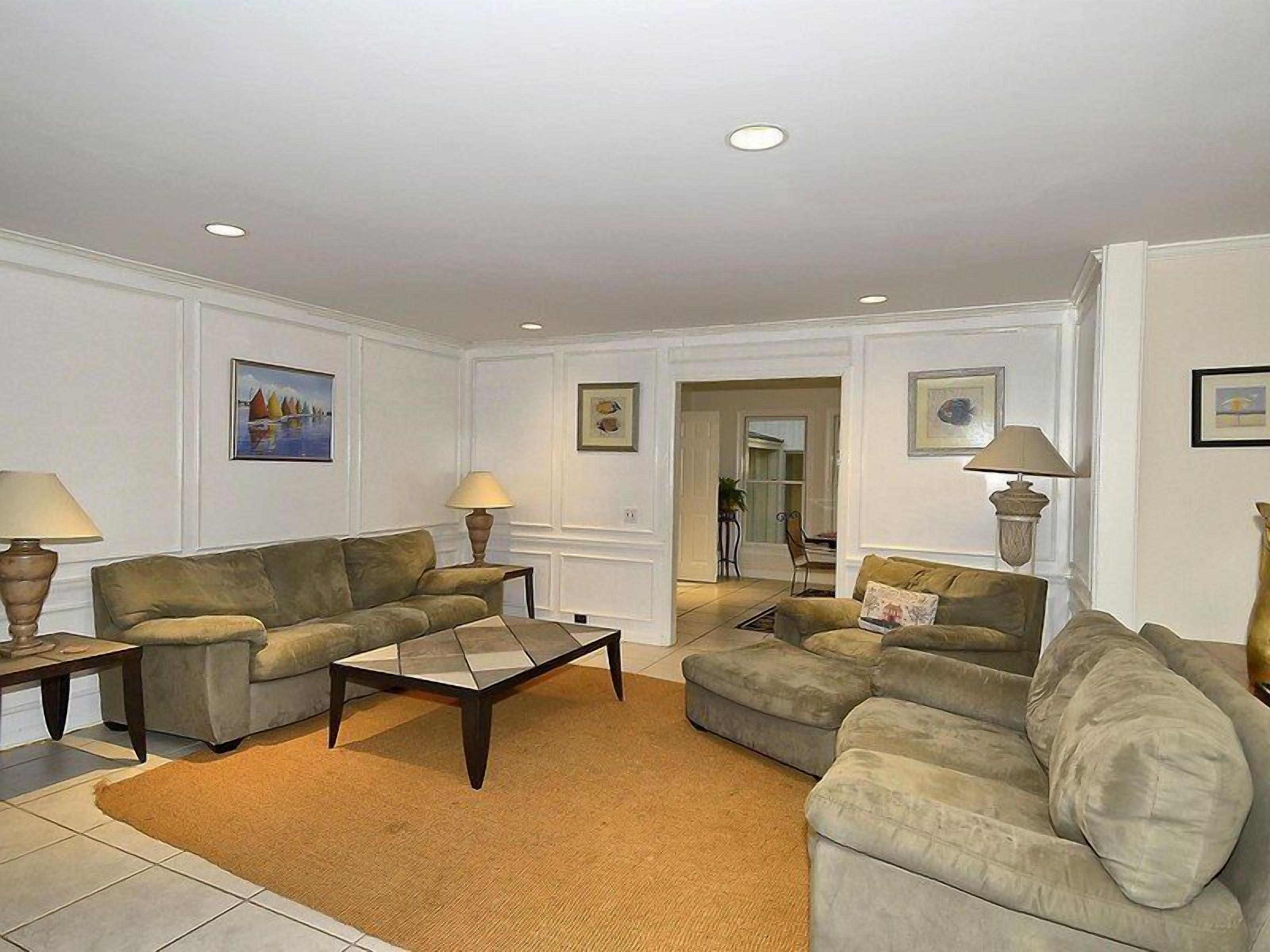 A living room with two sofas, a coffee table, and two lamps. There are framed pictures on the walls and a door leading to another room.