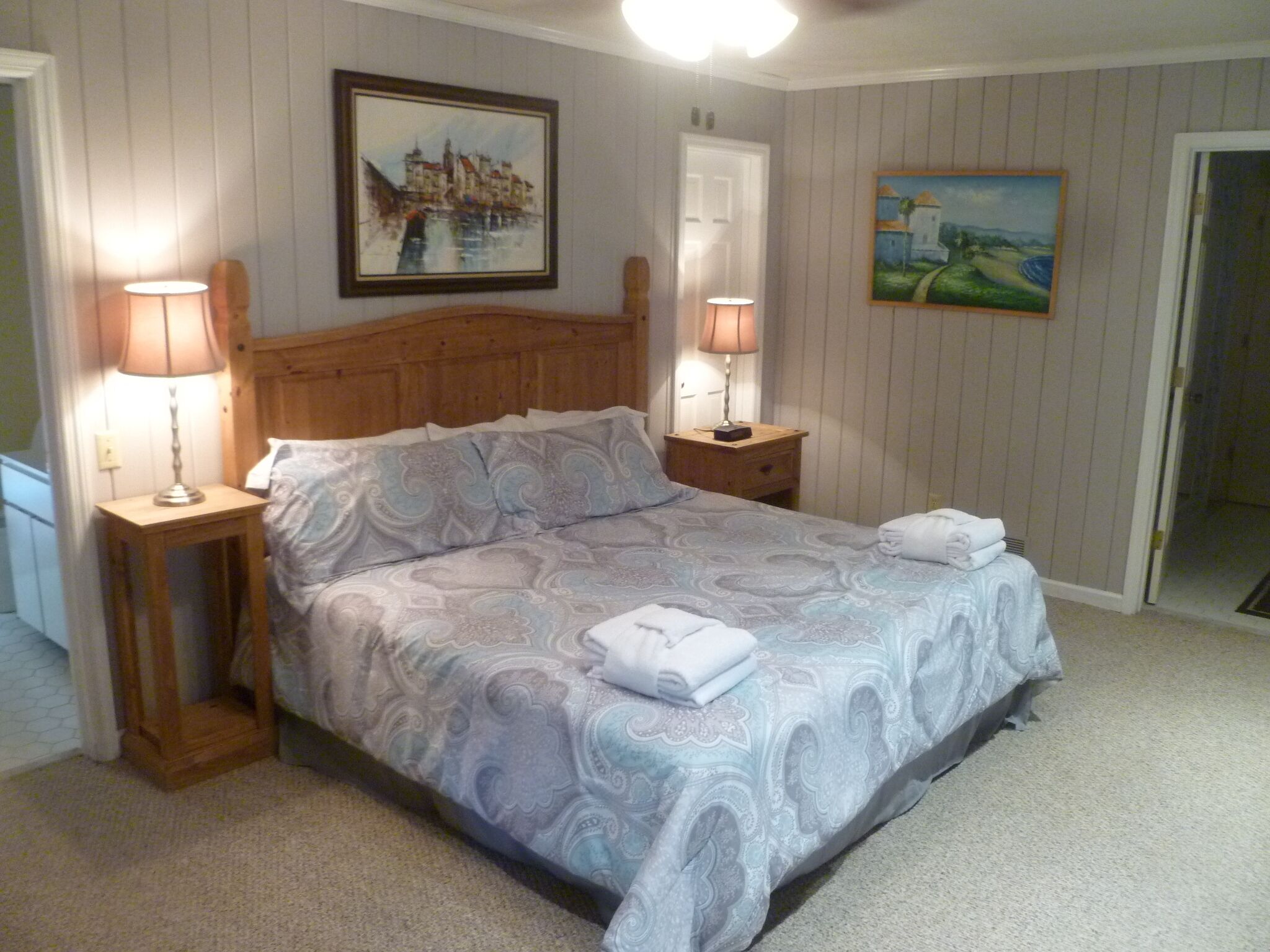 A bedroom with a wooden bed, two bedside tables with lamps, a painting on the wall, and a door leading to another room.