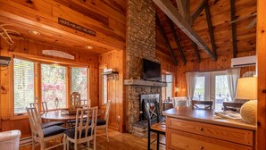 A cozy cabin interior with wooden walls, a stone fireplace, a dining area with chairs, and a kitchen counter. - Heather Heights Cottage - Cozy cabin with a View and Fire Pit, minutes to Boone! (Boone)