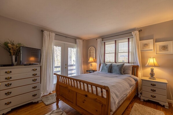 A bedroom with a wooden bed, a dresser, a television, and a window with curtains. - Heather Heights Cottage - Cozy cabin with a View and Fire Pit, minutes to Boone! (Boone)