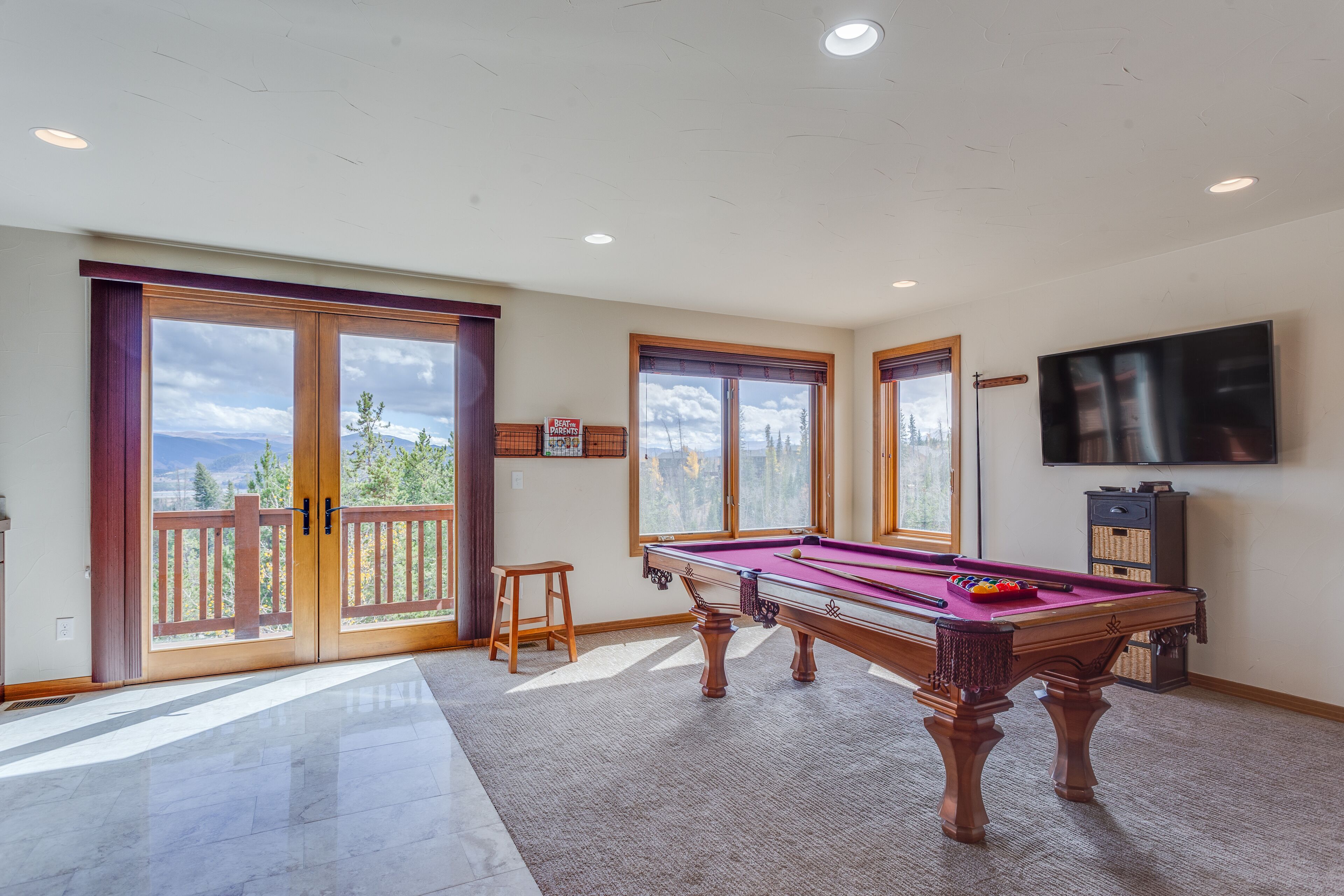 A pool table with a view of mountains through large windows, a television mounted on the wall, and a wooden deck outside.