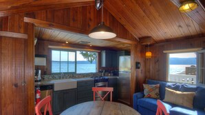 A cabin interior with a wooden ceiling, a dining table, a blue sofa, and a view of the lake through the windows. - Hood Canal, 1930's Beach Cottage,  315' Cove with Oysters! (Hoodsport)
