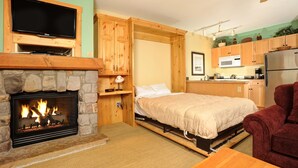 A compact room with a stone fireplace, a bed, a TV mounted above it, and a kitchenette with a microwave and refrigerator. - Fantastic Studio Condo Near the Lifts (Dillon)