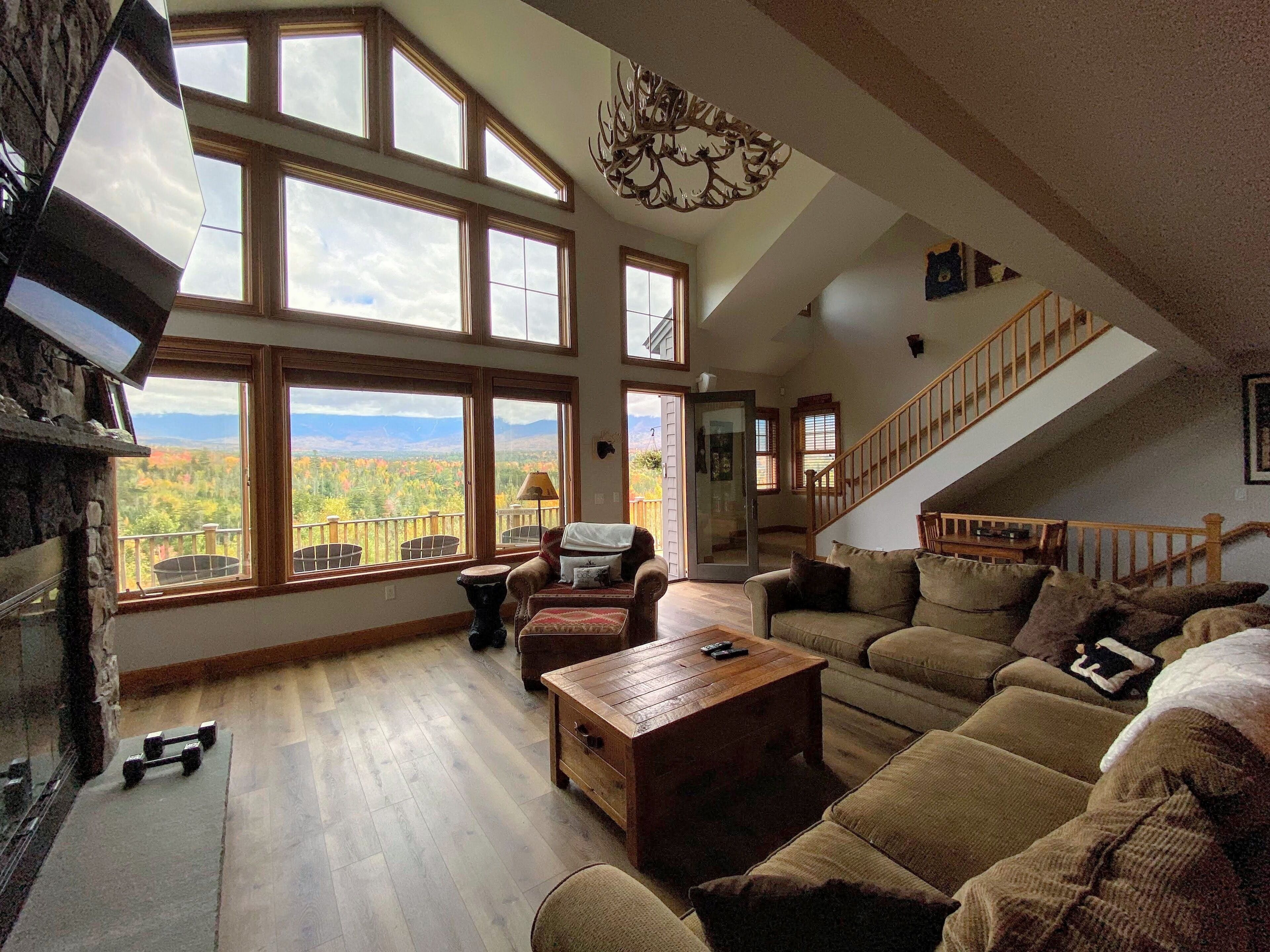 Un salón amplio con ventanas grandes que ofrecen vistas a la montaña, una chimenea y una escalera de madera.