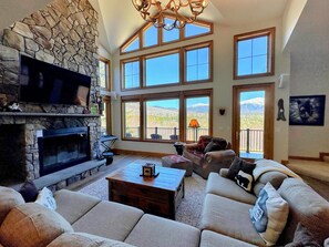 A spacious living room with a stone fireplace, large windows offering mountain views, and a cozy seating area.