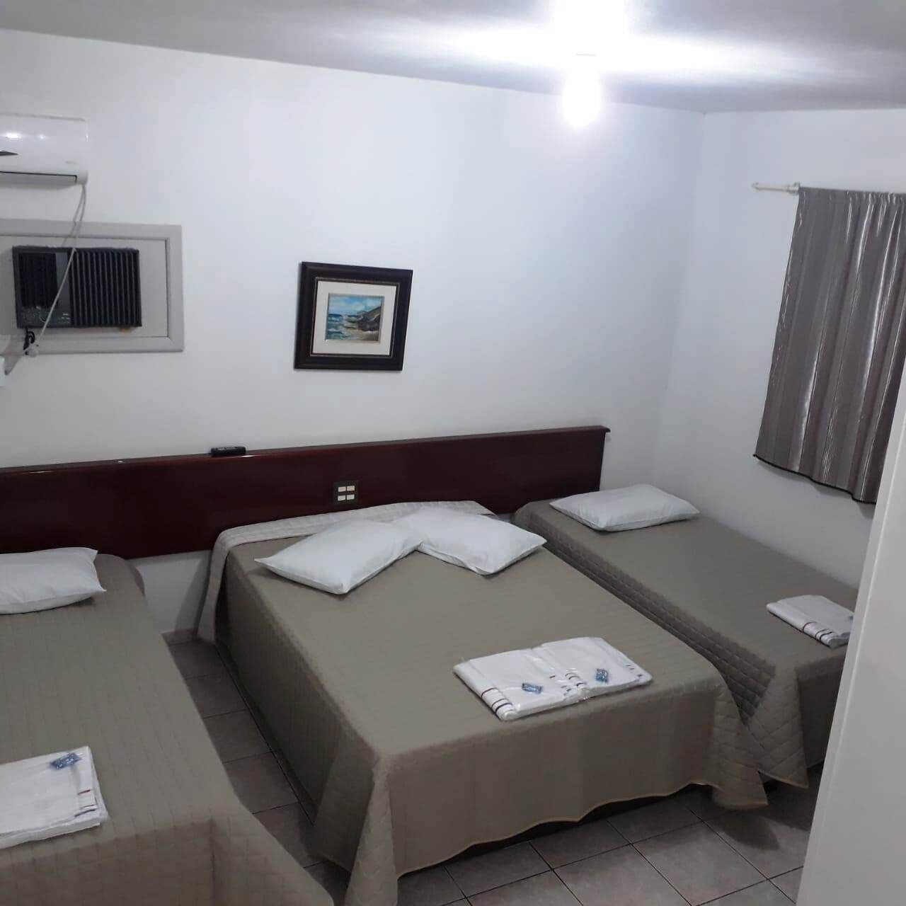 A hotel room with two single beds, a small table, a wall-mounted air conditioner, a framed picture, and a window with curtains.