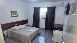 Ein Hotelzimmer mit einem Bett, einem kleinen Schreibtisch, einem Stuhl, einem Eimer und einem Fenster mit Vorhängen.