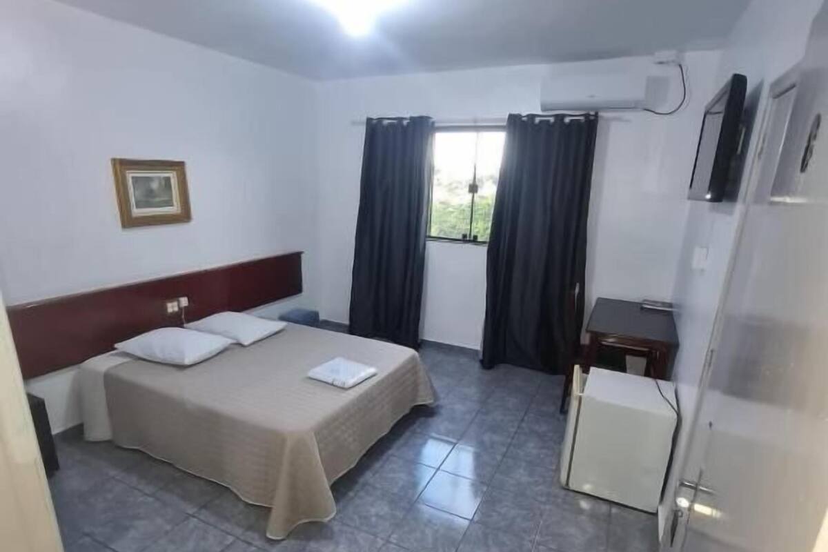 A hotel room with a bed, a small desk, a chair, a bucket, and a window with curtains.