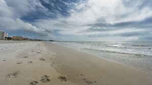 A beach with footprints leading to the water, a cloudy sky, and buildings in the background.