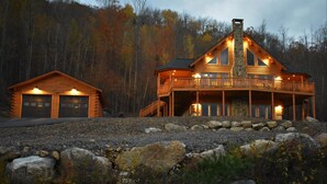 Un'ampia baita di legno con camino in pietra, un garage separato e un balcone affacciato su una zona boscosa.
