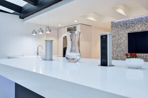 A modern kitchen with a white countertop, a unique glass vase, a built-in coffee machine, and a wall-mounted TV.