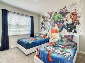 Un dormitorio con dos camas, una ventana con persianas, un mural de superhéroes en la pared y una mesita de noche con lámpara.