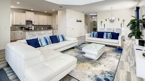 A modern living room with a white sectional sofa and a coffee table, a kitchen with white cabinets and a microwave, and a dining area with a white table and chairs.