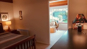 A hotel room with a bed, bedside table, lamp, and a view of the outdoor area through a doorway. - Peaceful Garden Retreat in Sandy Bay. (Sandy Bay)