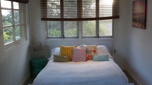 Een slaapkamer met een bed, kussens, een nachtkastje, een lamp en een raam met jaloezieën.