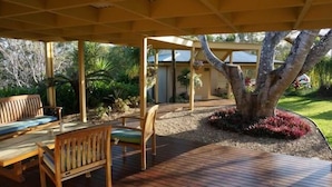 Een terras buiten met houten meubilair, een grote boom en een verzorgde tuin.