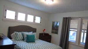 Quarto com cama, mesa de cabeceira, quadro emoldurado e janela com cortinas.