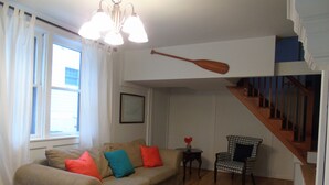 Uma sala de estar com um sofá bege, almofadas coloridas, um tapete estampado, uma pá de madeira, uma cadeira e uma escada.