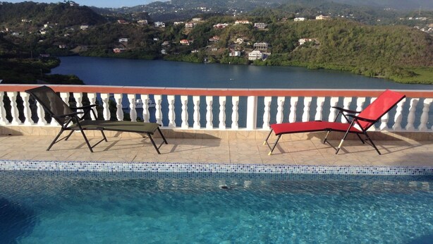 A swimming pool with two lounge chairs, a tiled deck, and a view of a lake and hills in the background. - Paradise on the Isle of Spice (Calivigny)