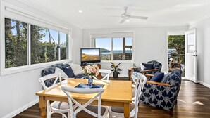A spacious room with a wooden dining table, white chairs, a sofa with patterned cushions, and a television displaying a scenic view. - Jetty Splendour Apartment (Coffs Harbour)