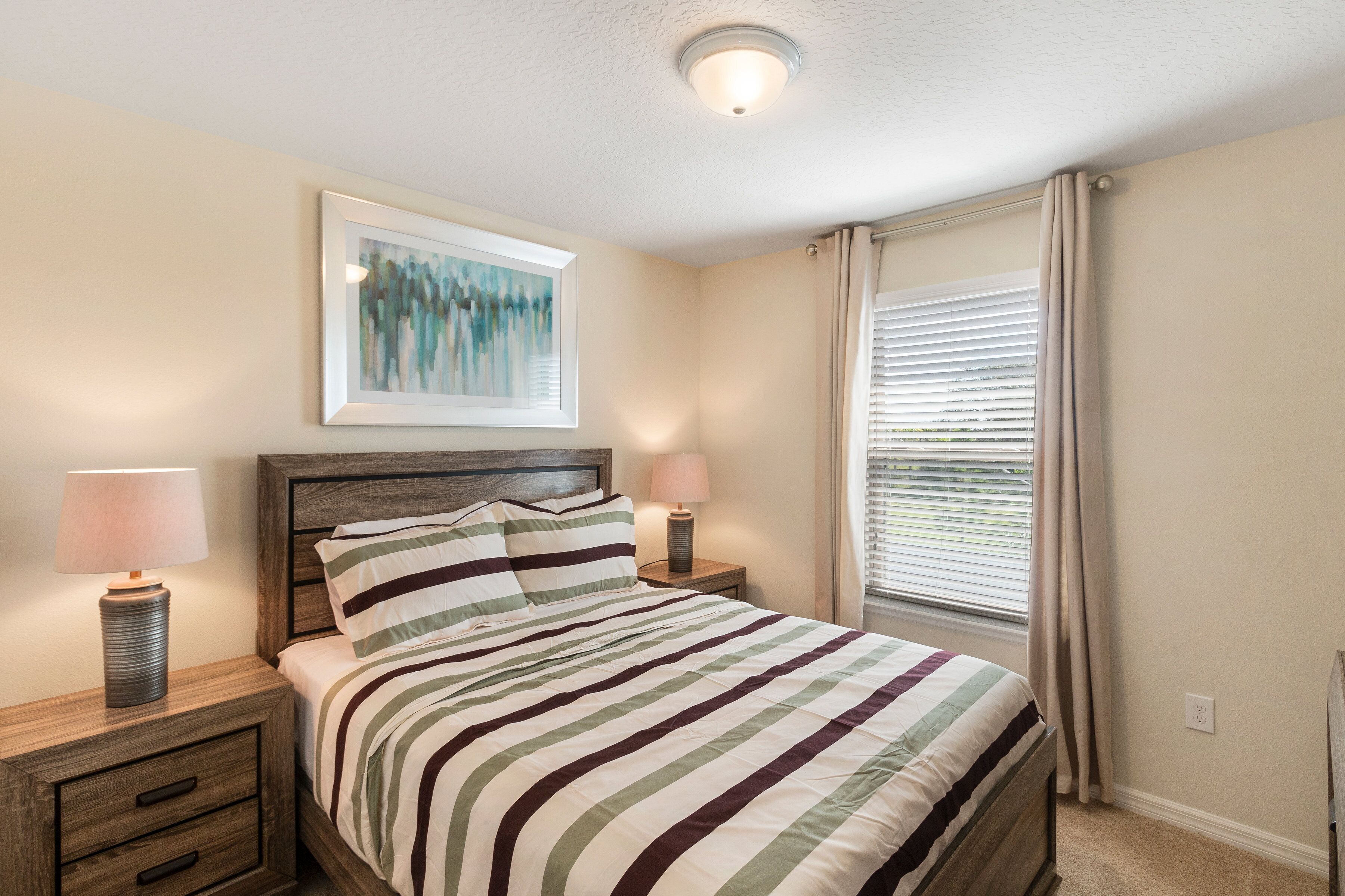 A bedroom with a bed, two bedside tables with lamps, a framed artwork on the wall, and a window with curtains.