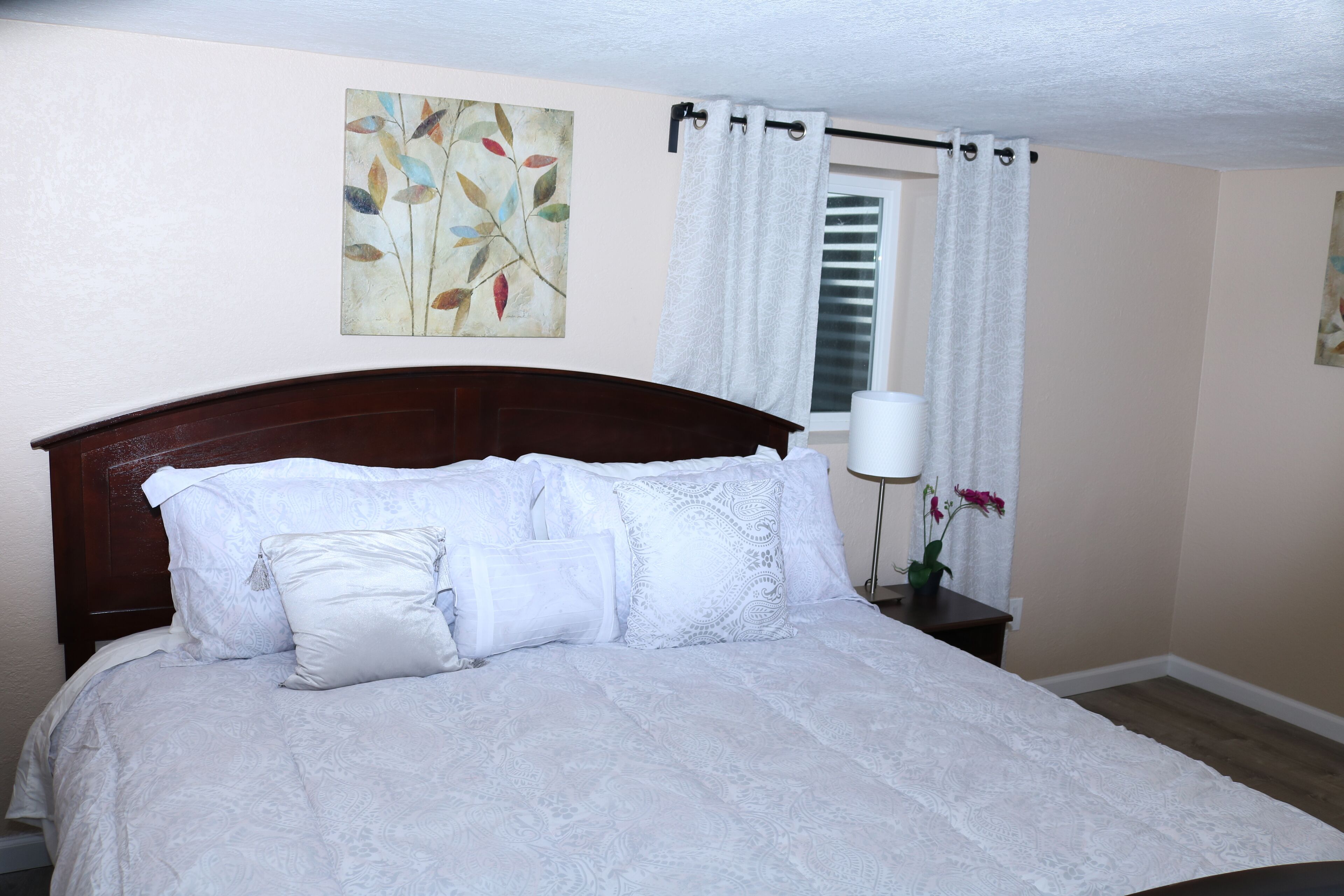A neatly made bed with white linens and a wooden headboard in a room with cream walls and a window with white curtains.