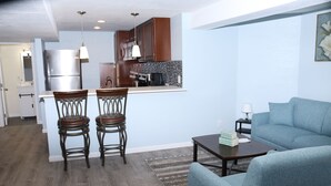 Une cuisine moderne avec des armoires en bois, un réfrigérateur en acier inoxydable et un four à micro-ondes encastré. On y trouve un coin repas avec deux chaises en bois et une petite table. Un canapé bleu se trouve dans l’espace salon, et une lampe est posée sur une table d’appoint.