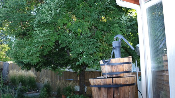 Une fontaine en forme de tonneau en bois, située dans un jardin agrémenté d’une clôture en bois et de diverses plantes.