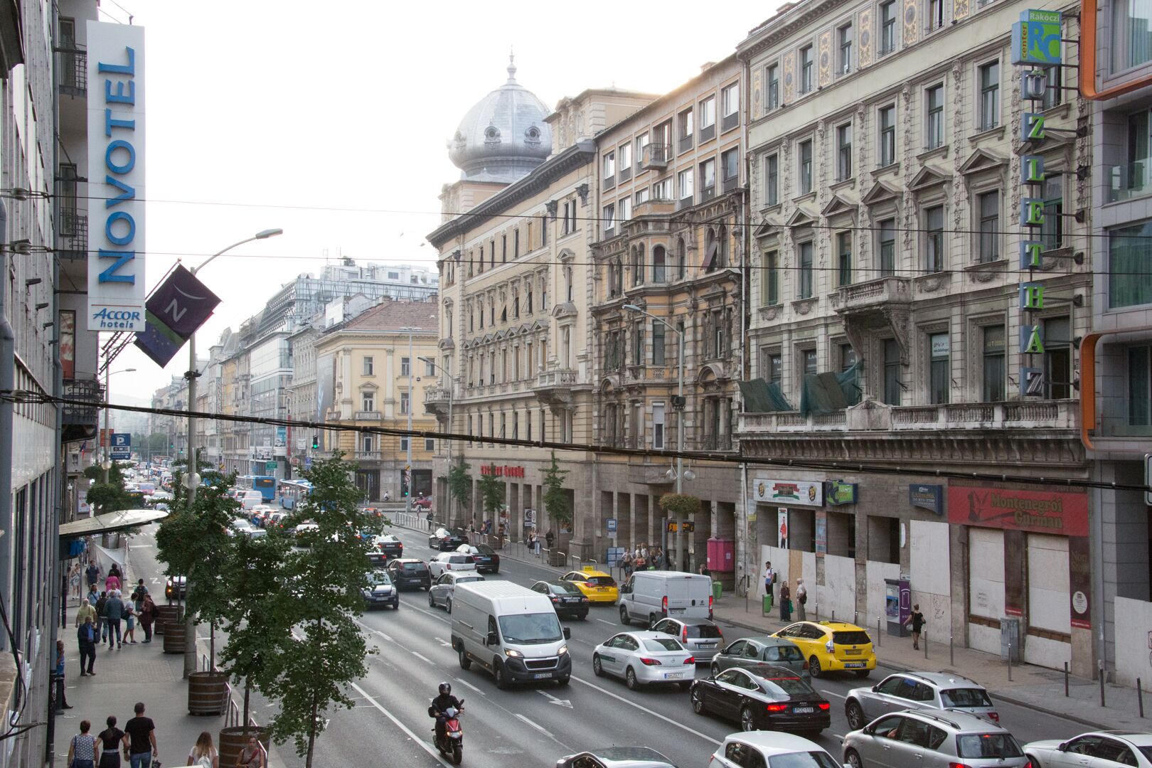 Eine belebte Stadtstraße mit historischen Gebäuden, einem Novotel Hotelschild und einer Straßenbahnlinie.