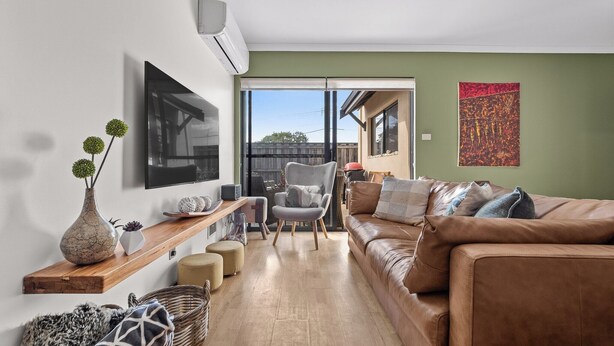 A living room with a brown leather sofa, a grey armchair, a wooden shelf with decorative items, and a large TV on the wall.