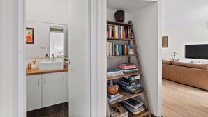 A modern bathroom with a white sink, mirror, and cabinets. Adjacent is a bookshelf with various books and decorative items.