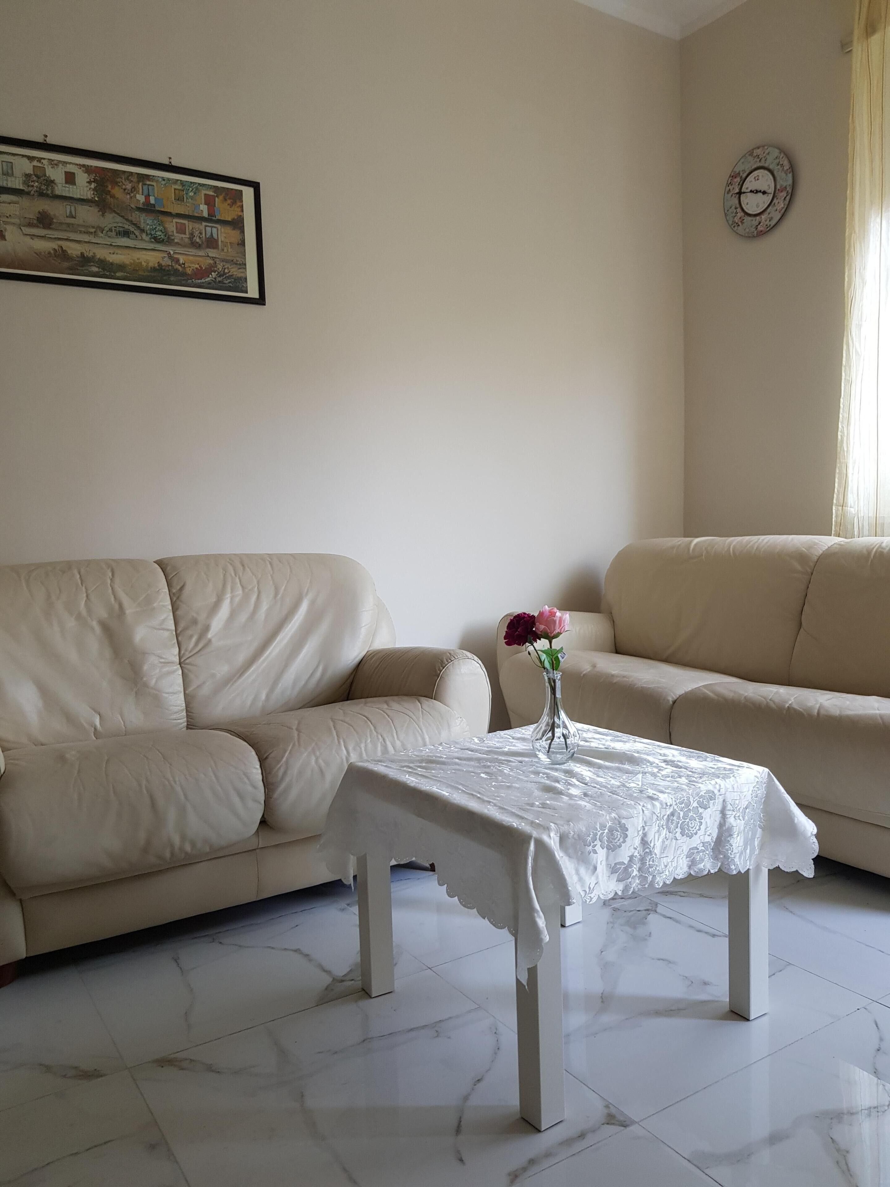 A living room with two beige sofas, a white table with a lace tablecloth, and a vase with flowers.