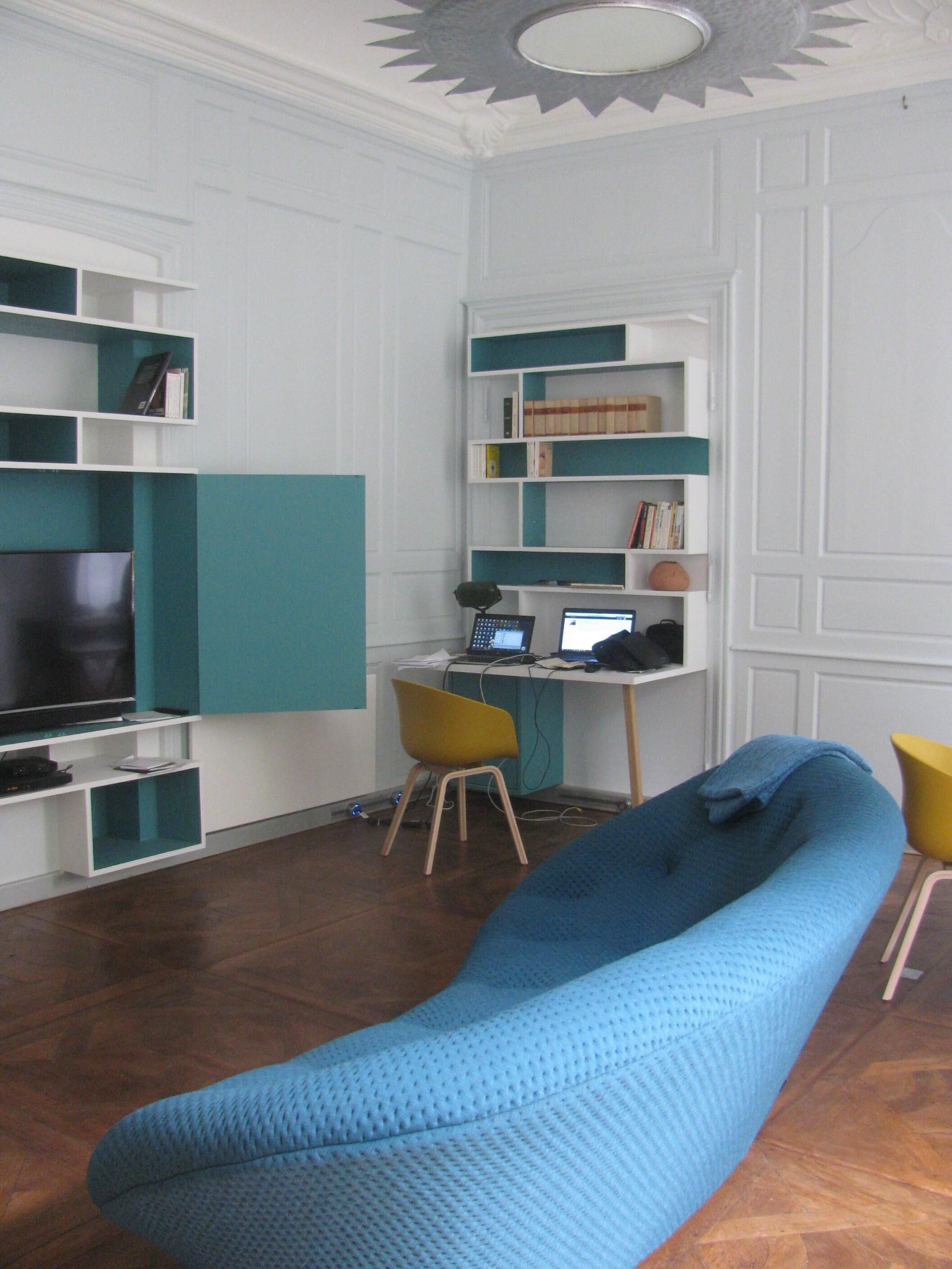Een woonkamer met een blauwe bank, een gele fauteuil, een televisie en een bureau met een computer.