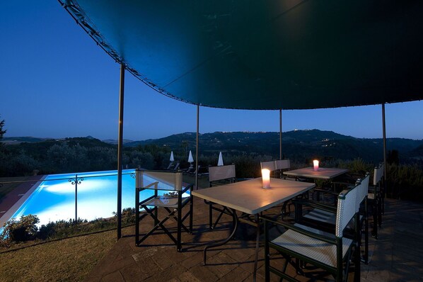 Un patio exterior con piscina, mesas y sillas bajo una marquesina, con vistas a un paisaje montañoso.