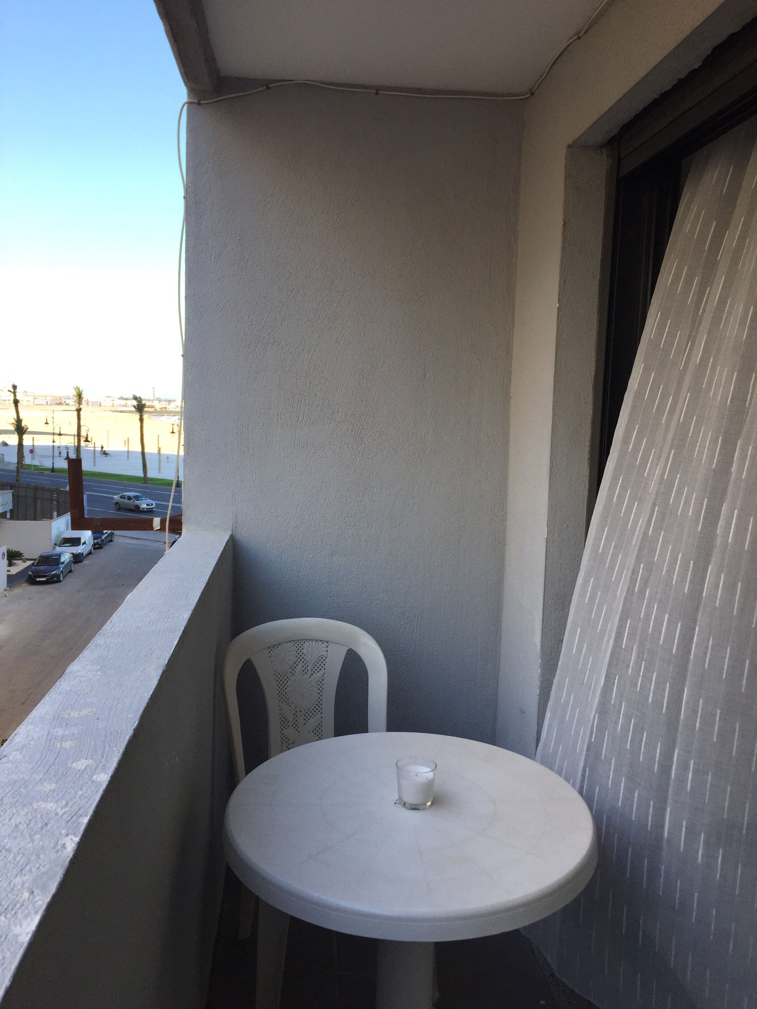 A balcony with a small round table and a white chair, overlooking a street with parked cars and palm trees.