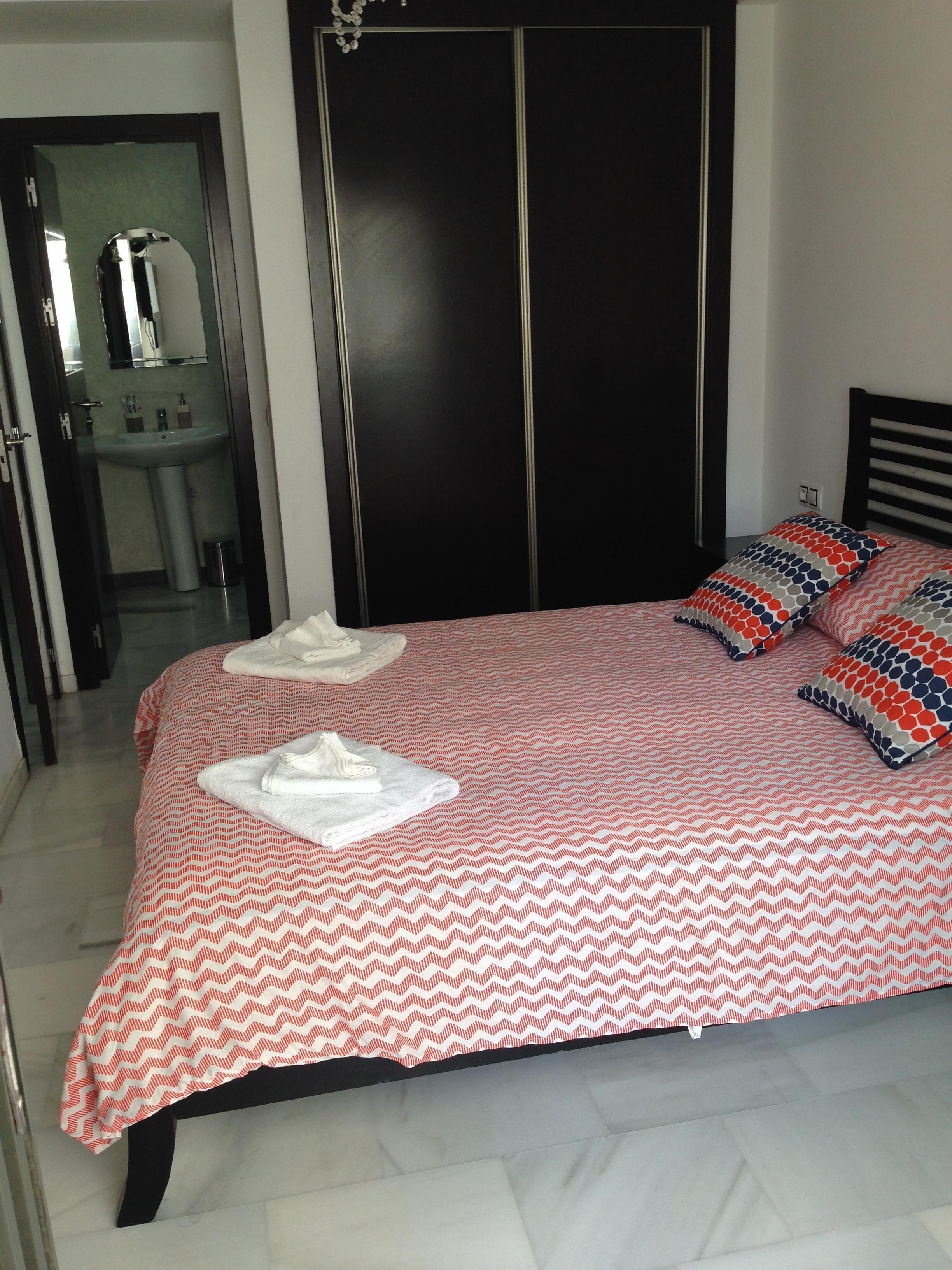 Une chambre à coucher avec un lit, des oreillers, une table de chevet, un miroir et une salle de bain visible au fond.