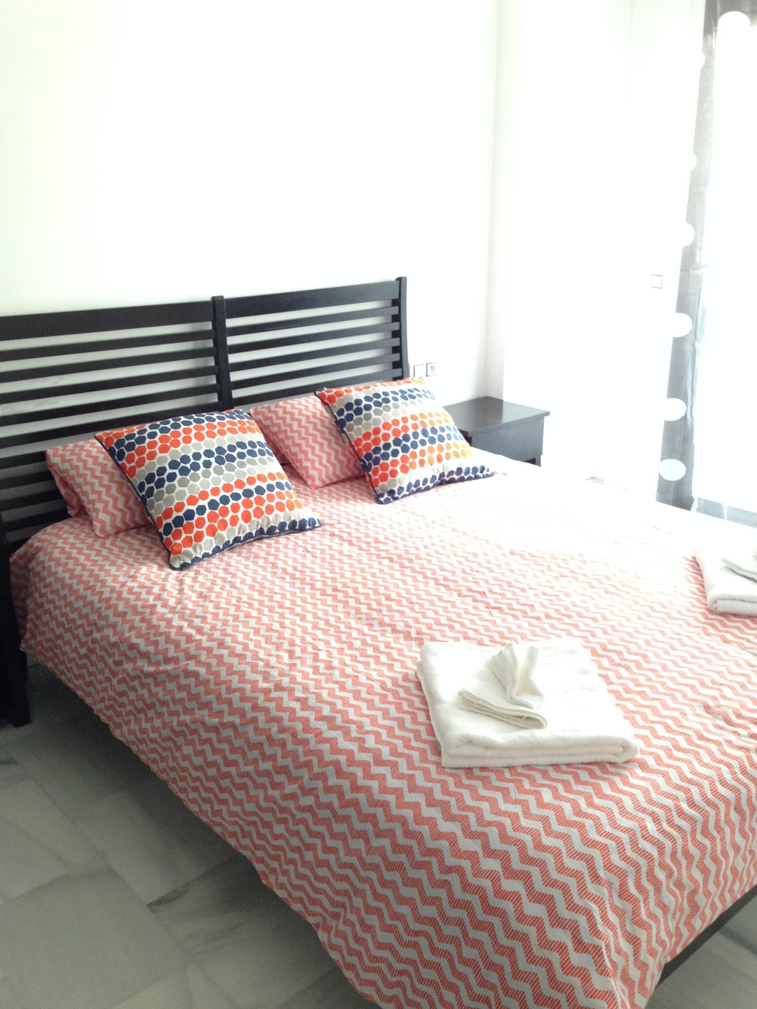 Een net opgemaakt bed met een bedspread met patroon en kussens, een zwart hoofdbord, een nachtkastje met een lamp en een raam met gordijnen.