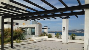 Een buitenterras met een pergola, uitzicht op zee en een gebouw met een glazen gevel.