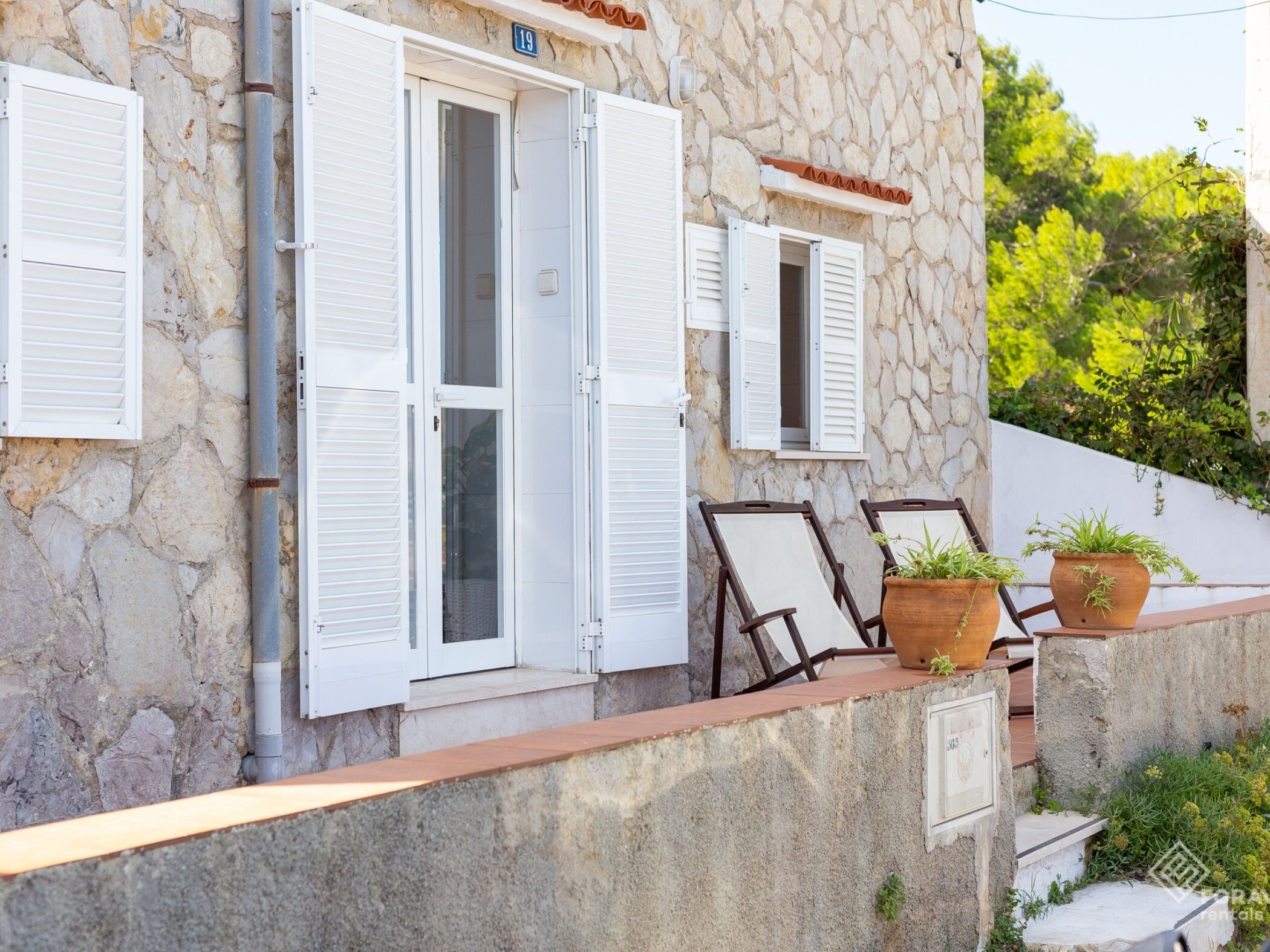 A stone building with white shutters, a number '19' sign, and outdoor seating with potted plants.
