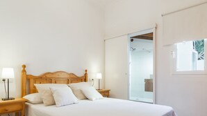Un dormitorio con una cama de madera, dos mesitas de noche con lámparas, un ventanal grande y una puerta corredera de vidrio que da a un baño.