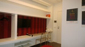 Habitació amb un televisor muntat sobre una paret vermell rayat, una banca blanca i una porta amb un quadre d'un edifici.