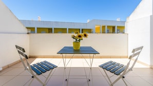 Ein Balkon mit Tisch und zwei Stühlen, ein gelber Topf mit Sonnenblumen und im Hintergrund ein Gebäude mit gelben Türen.
